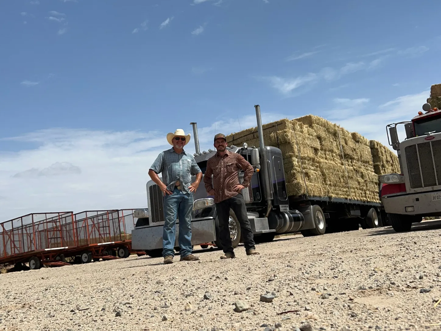 Two men standing in front of a hay-loaded semi truck.