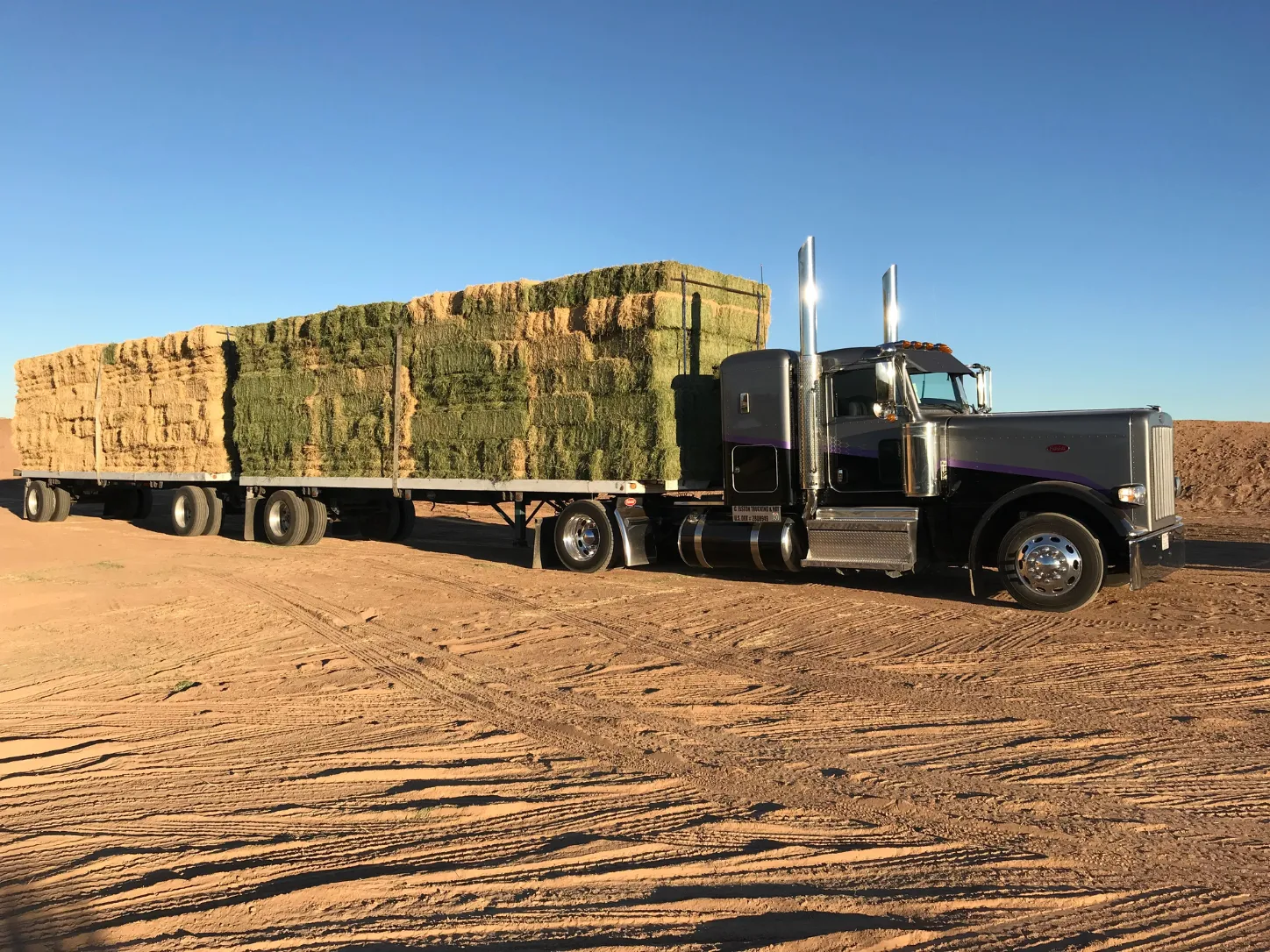 Semi truck loaded with stacked hay bales on a dirt field.